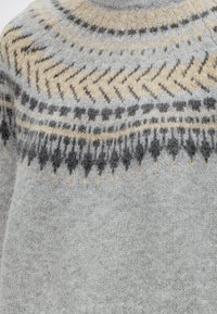 Gros plan d'un pull en maille gris avec un motif rond à l'empiècement en beige, gris foncé et gris clair avec des motifs géométriques.