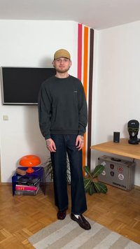 Ciemnozielona bluza, granatowe jeansy z szerokimi nogawkami, brązowe buty i beżowa czapka. Charakterystyczne elementy to tatuaże i kolorowa ściana w tle.