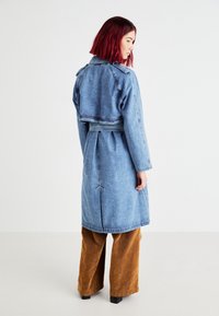 Cappotto lungo in denim con retro corto, vita cintrata e maniche lunghe. Caratterizzato da un lavaggio blu e uno spacco centrale sul retro. Indossato sopra pantaloni larghi marroni.