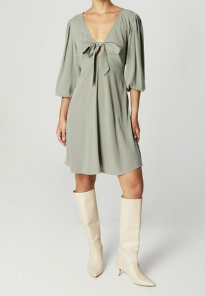 Femme portant une robe longueur genou vert sauge avec des manches bouffantes nouées sur la poitrine, associée à des bottes à talons montantes jusqu'au genou de couleur blanc cassé.