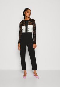 Čierny overal s dlhými čipkovými rukávmi, biely korzet s čipkovými detailmi a priliehavé čierne nohavice. Rúžové lodičky dopĺňajú outfit.