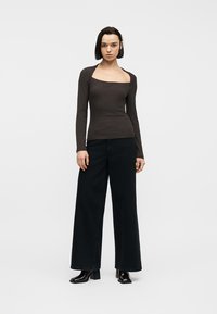 Νέα γυναίκα που φοράει καφέ μπλούζα με τετράγωνο λαιμό και μακριά μανίκια, μαύρο παντελόνι με φαρδιά πόδια και μαύρες μπότες με τακούνι, στέκεται μπροστά σε απλό λευκό φόντο.