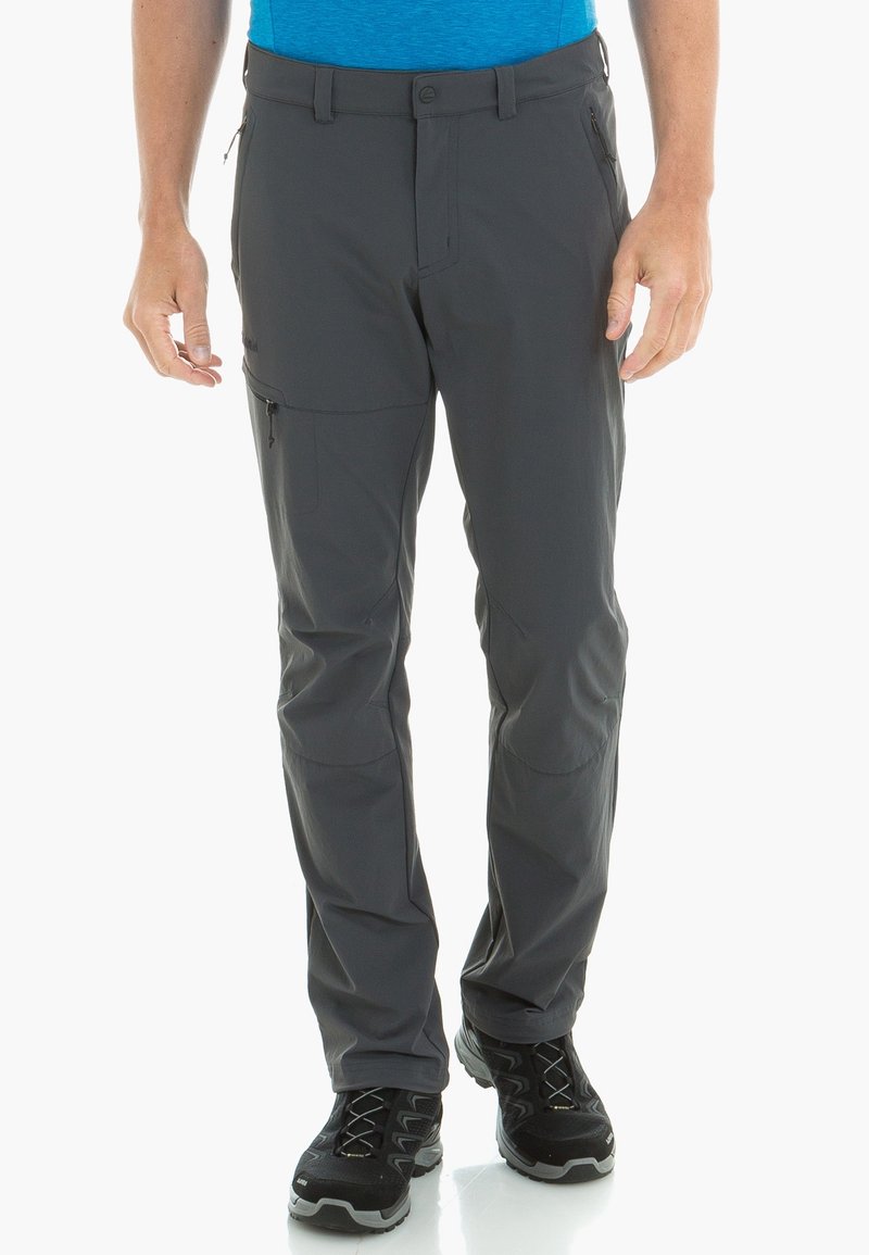 Homme portant un pantalon de randonnée gris foncé avec une poche cargo zippée et des chaussures de randonnée noires, debout sur fond blanc.