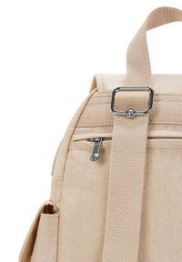 Zaino in tessuto beige con tasca con zip, una tracolla texturizzata e un accento in metallo argento sul punto di attacco della tracolla.