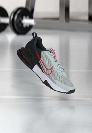 Ανοιχτό γκρι και μαύρο sneaker Nike Air Max με κόκκινες λεπτομέρειες που αιωρείται πάνω από γυαλιστερό πάτωμα σε χώρο γυμναστηρίου με βάρη στο φόντο.