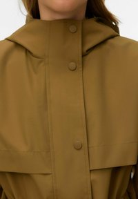 Gros plan sur une personne portant un imperméable jaune moutarde à capuche avec trois boutons-pression sur le devant.