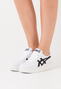 Baskets blanches Asics avec des rayures noires portées pieds nus sur un fond uni, montrant les jambes inférieures et les chaussures.