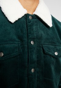 Veste en velours côtelé vert foncé avec boutons-pression argentés et col sherpa blanc, montrant le haut de la poitrine et l'encolure.