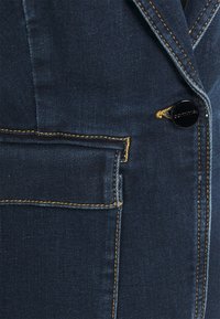 Blazer en denim foncé avec coutures jaunes, une poche verticale et un bouton en métal rond avec un détail de logo gravé.