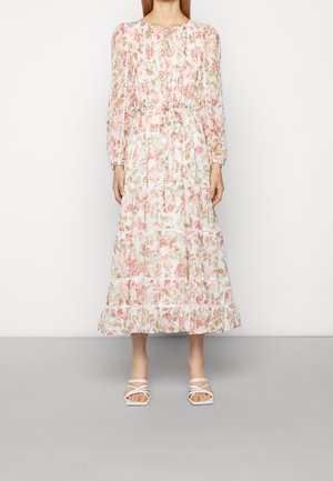 Kvinde iført en lang blomsterkjole med pink og grønne mønstre, lange ærmer, en indsnævret talje og hvide hælede sandaler med stropper.