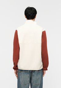 Άνδρας που φοράει λευκό αμάνικο γούνινο γιλέκο πάνω από μπλούζα μακρυμάνικη σκουριάς χρώματος και άνετο μπλε τζιν, στέκεται με την πλάτη προς τα εμπρός σε απλό φόντο.