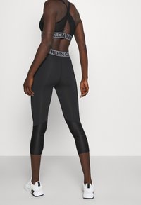 Zwarte sport bh met gaaspanelen en elastische band, gecombineerd met zwarte capri leggings met een glanzende afwerking en contrasterende texturen.