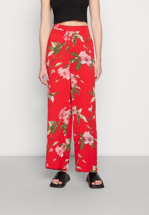 Calças largas vermelhas com um padrão floral, apresentando flores cor-de-rosa e folhas verdes, feitas de um tecido leve, combinadas com sandálias pretas.
