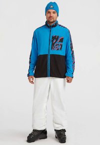 Blue and black zip-up jacket with a patterned pocket, paired with white pants and black ski boots. Features a blue knit beanie.