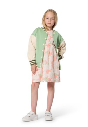 Mintgroene en crèmekleurige bomberjack met gestreepte manchetten over een jurk met bloemenpatroon in pastelkleuren; witte sneakers maken de outfit compleet.