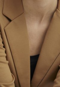 Blazer marrón con una textura suave y solapas estructuradas, usado sobre una parte superior oscura. Primer plano que muestra el detalle del escote y el collar.