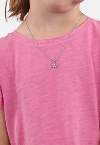 Collana a forma di stella argentata con un accento blu, indossata sopra una maglietta rosa e testurizzata. La catena è delicata e presenta una chiusura semplice.