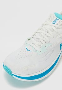 Chaussure de sport blanche avec une tige en mesh légère, des accents turquoise et une semelle texturée. Présente des lacets et des éléments de design de marque sur le côté.