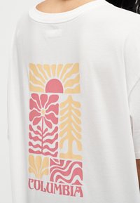 Λευκό T-shirt με ροζ και κίτρινο αφηρημένο γραφικό λουλουδιών και ήλιου και το κείμενο "COLUMBIA" στην πλάτη, που φοριέται από άτομο.