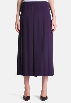 Mujer con una falda midi plisada de cintura alta en color púrpura oscuro combinada con tacones negros de punta afilada, de pie frente a un fondo liso.