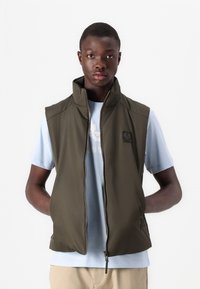 Olive green sleeveless vest with a high collar, zip front, and subtle logo patch, layered over a light blue t-shirt. Smooth texture.