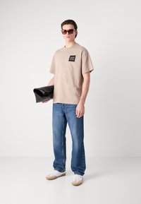 T-shirt bej cu maneci scurte, cu o dungă în față, blugi din denim albastri și adidași albi cu accente bej. Ținând o poșetă neagră.