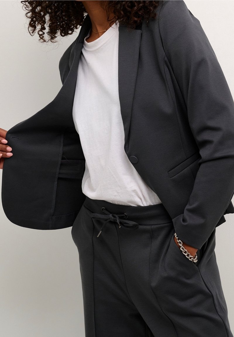 Blazer gris oscuro con un solo botón, que incluye un bolsillo interior. Capas sobre una camisa blanca, llevado con pantalones gris oscuro a juego.