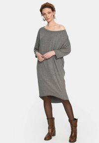 Szary, oversize sukienka z dekoltem łódka, długimi rękawami i zaokrąglonym dołem. Noszona z brązowymi wysokimi kozakami i półprzezroczystymi brązowymi rajstopami.