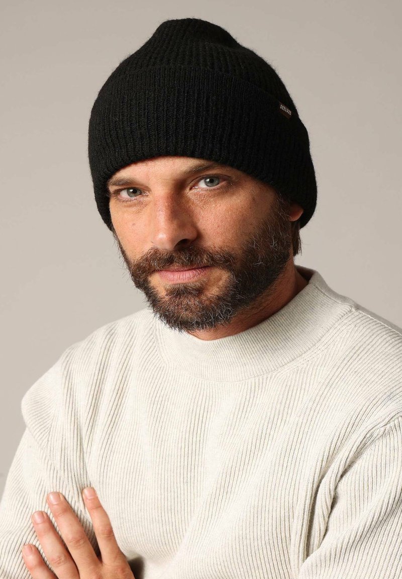 Zwarte gebreide beanie met een geribbeld textuur, voorzien van een klein merkembleem. Draagt een lichtgrijze, geribbelde trui met hoge hals.