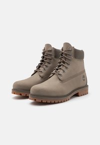 Botas de tobillo impermeables color beige con parte superior de cuero suave, suela de goma y cordones redondos. Presenta un detalle de logo en el lateral.