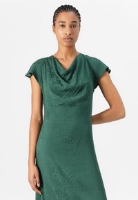 Green dress with a cowl neck, short sleeves, textured fabric featuring a subtle dot pattern. Fitted silhouette with an elegant drape.