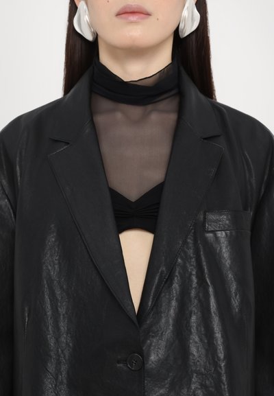 Blazer din imitație de piele neagră, lucioasă, cu un singur nasture, având o decupare adâncă în V și buzunar. Sub el, un top transparent cu gât înalt și un sutien negru.