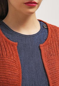 Cardigan aperto color ruggine in maglia a trama larga sopra un top in denim liscio di colore blu scuro. Presenta un collo rotondo e un'acca di bottone sulla spalla.
