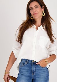 Vrouw in een witte blouse met knopen en een blauwe spijkerbroek, geaccessoireerd met gouden oorringen en armbanden, tegen een effen achtergrond.