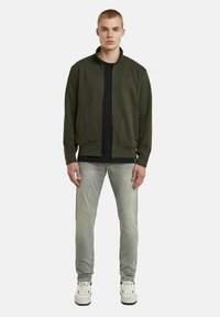 Giacca nera oliva con zip e colletto alto, abbinata a una maglietta nera e jeans grigio chiaro. Le scarpe sono bianche con dettagli neri.