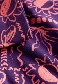 Lila Stoff mit orangenen abstrakten Blumen- und geometrischen Mustern, zeigt eine umgeklappte Ecke und strukturierte Nähte.