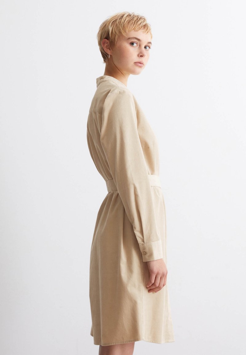 Beige knielanges Kleid mit langen Ärmeln, einem knöpfbaren Kragen und einem Stoffgürtel in der Taille. Glatte Textur und schlichtes Design.