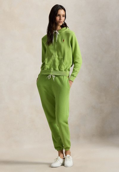 Πράσινο φούτερ με φερμουάρ, μπροστινή τσέπη και κορδόνια, σετ με ταιριαστό παντελόνι φόρμας με ελαστικές μανσέτες και απλά λευκά αθλητικά παπούτσια.
