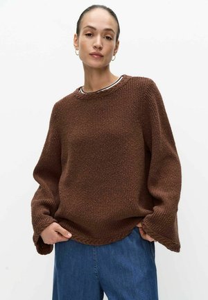 Vrouw met achterovergekamd haar, oorbellen in de vorm van ringen, een losse bruine gebreide trui en een blauwe spijkerbroek, staand tegen een witte achtergrond.