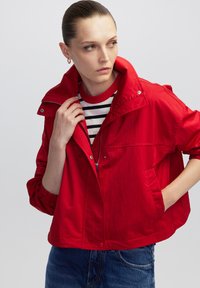 Vrouw met slicked-back haar, draagt grote oorbellen, een rode zip-up jas over een gestreept shirt en blauwe jeans, kijkend naar haar linkerzijde.