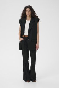 Must kohandatud vest õlapadjadega, mis on kombineeritud kitsaste mustade pükstega. Omab vöö, tasku detaili ja teravat pruuni jalanõud.