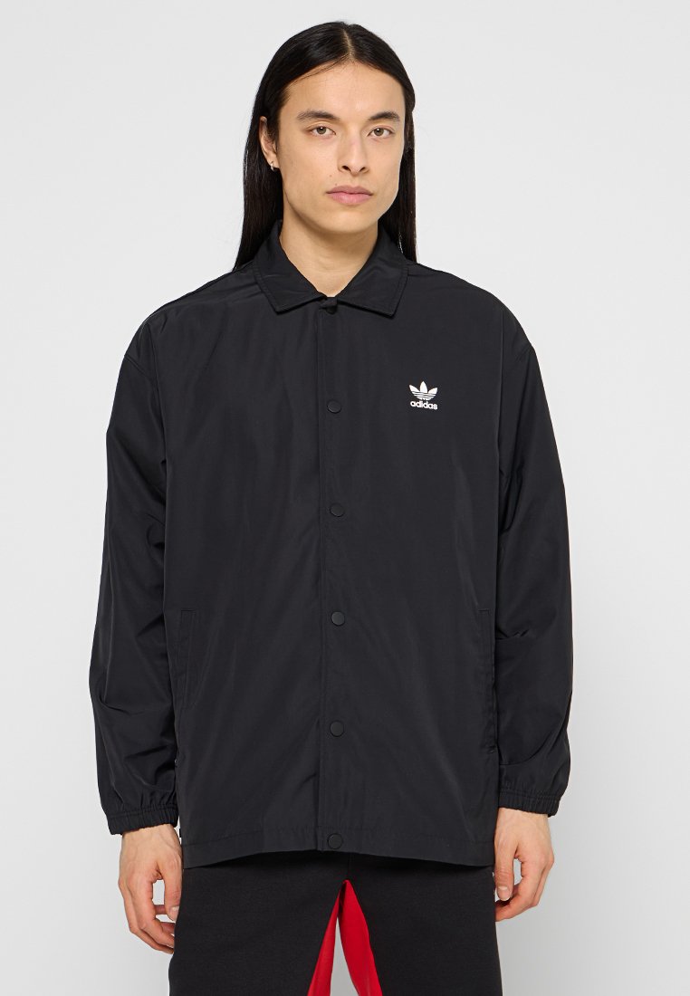 Schwarze Windjacke mit Kragen, Druckknöpfen, Fronttaschen und weißem Adidas-Logo auf der linken Brust.
