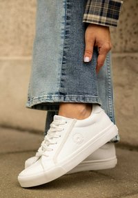 Białe sneakersy wykonane z gładkiej skóry z teksturowanymi gumowymi podeszwami. Wśród cech znajdują się sześć oczek, subtelne logo i płaskie sznurowadła; zestawione z dżinsami.