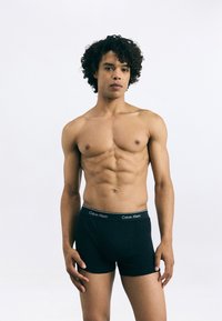 Jauns vīrietis ar cirtainiem matiem, tērpies melnās Calvin Klein bokseršortos, stāv uz vienkārša balta fona.