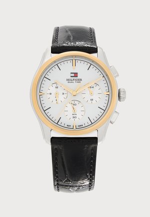 Round silver and gold Tommy Hilfiger dual time watch with white dial, three subdials, gold hands, and a black leather strap.