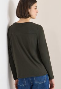 Olive green long-sleeve shirt with a round neckline and shoulder seam detailing, paired with blue jeans. Fabric is soft and smooth.