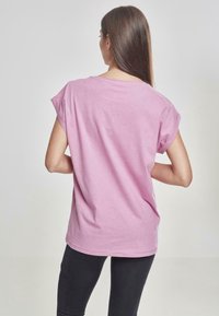 Camiseta de algodón rosa con mangas cortas enrolladas, corte holgado y dobladillo redondeado. Mostrada desde atrás con un fondo neutro.