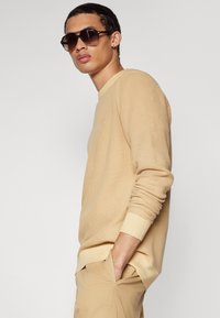 Beige strukturiertes Langarmhemd mit gerippten Bündchen. Das Model trägt eine dunkle Sonnenbrille und präsentiert ein neutrales und geschichtetes Outfit mit hellen Hosen.