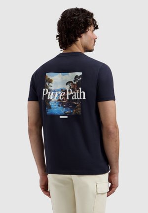 Man draagt een marineblauw T-shirt met een schilderachtige rivier en bomen op de achterkant, met de tekst "Pure Path" eroverheen en een klein label eronder.