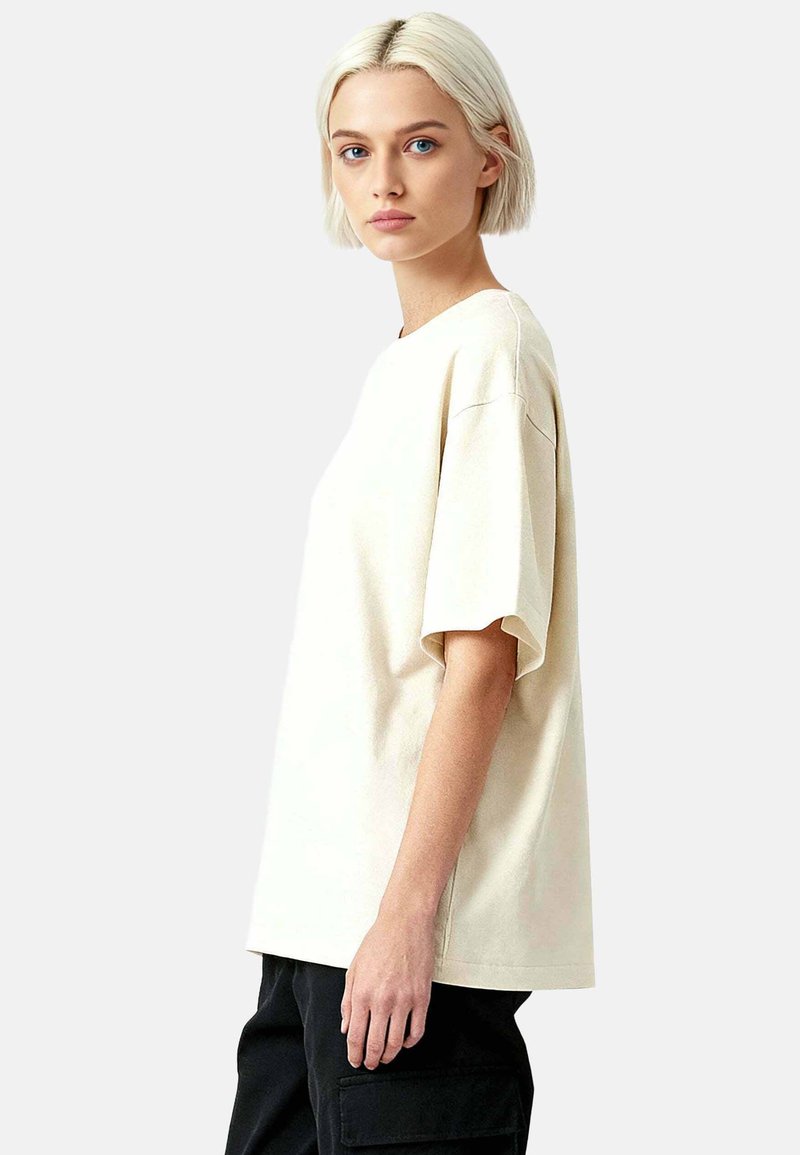 Jeune femme aux cheveux courts platine portant un t-shirt crème surdimensionné et un pantalon noir, debout devant un fond gris uni.